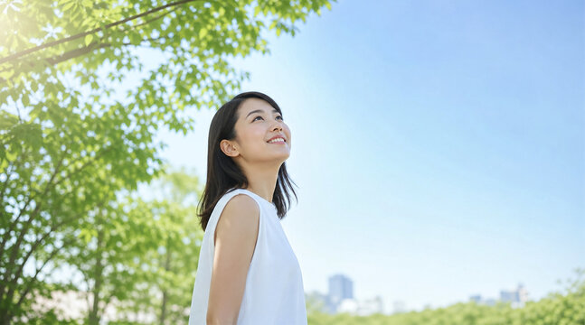 新緑の木々の下から青空を見上げて微笑む爽やかな若い日本人女性