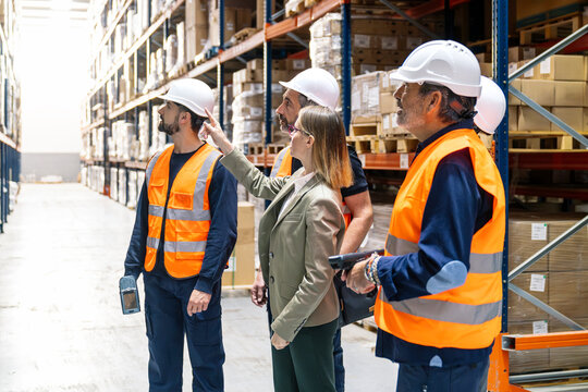Team of logistics professionals with hard hats and safety vests conducting an inspection and discussing operations in a busy warehouse