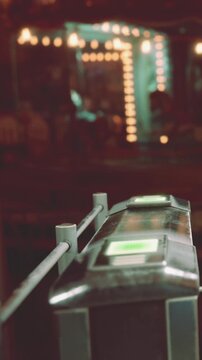 A turnstile stands in the foreground as people gather in a busy venue at night. Bright lights from the background create a lively atmosphere. The excitement is evident as guests enter.