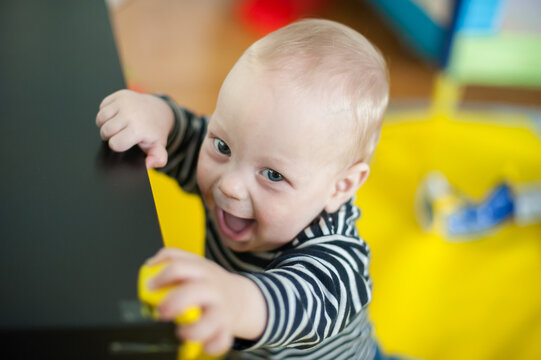 Cheerful baby boy holding onto a black table in a bright home living room while smiling with pure joy during a playful morning exploration at home