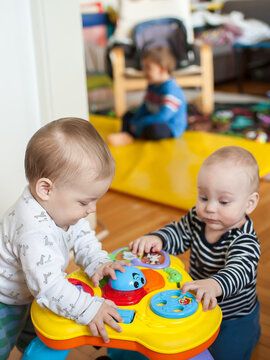 Three young brothers toddlers gather around a colorful activity table to press buttons and manipulate toys while a sibling sits on a yellow floor mat in the background 