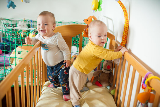 Two happy little brothers stand side by side holding onto the wooden railings of their crib while surrounded by soft plush animals and hanging rattles during a cheerful morning activity at home