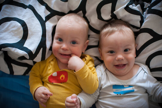 Two adorable twin infants with sparkling eyes lay together on soft linens, sharing joyous laughter and sweet expressions during a peaceful morning rest in their warm bedroom