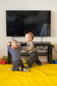 Twin toddlers curiously explore a bright home near a sleek black TV console on a sunny afternoon