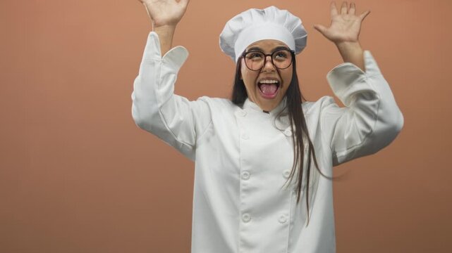 Young female chef with toque and glasses raises fists and hands to face in surprise then celebration in a studio; joy success triumph.