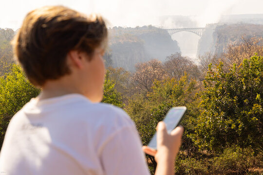 young boy looking at smart phone overlooking Victoria Falls, Zimbabwe, Africa