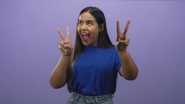 Young woman shows both hands in double v peace sign and puckered lips against purple studio backdrop; playful confidence.