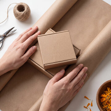 Hands wrapping a stack of plain brown kraft boxes in kraft paper with twine and marigold petals