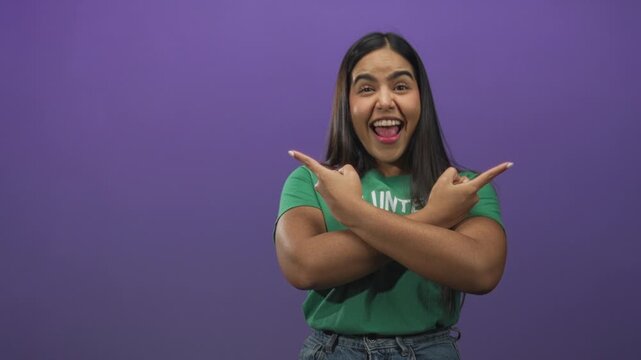 Woman wearing green volunteer tshirt crosses arms and points both index fingers to sides in studio against purple backdrop; community service joy.