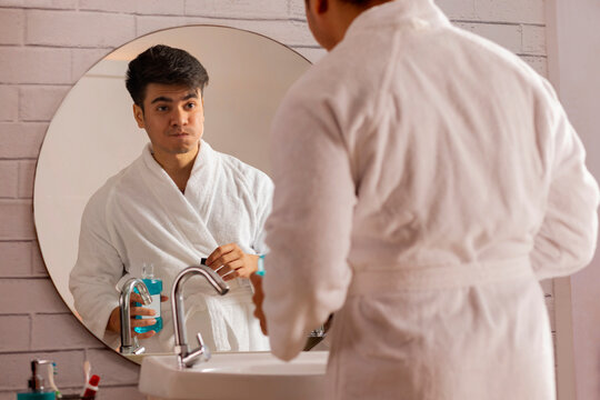 Young man in bathrobes gargling with mouthwash in front of bathroom mirror