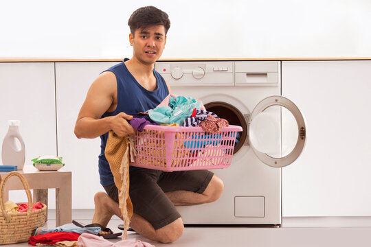 Smiling young man sitting in front of washing machine with laundry basket