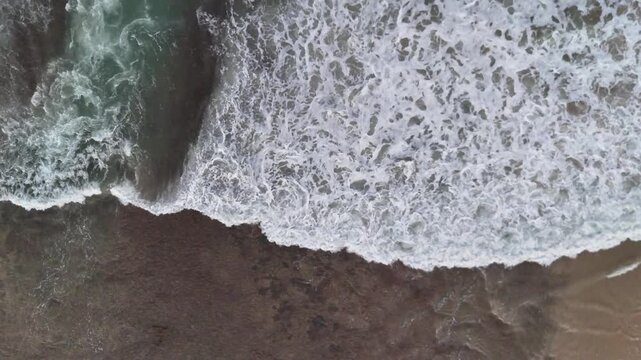 Aerial drone footage of foamy white waves moving toward the beach, colliding with dark coastal rocks. Captures the raw beauty of sea, surf, and shoreline in motion.