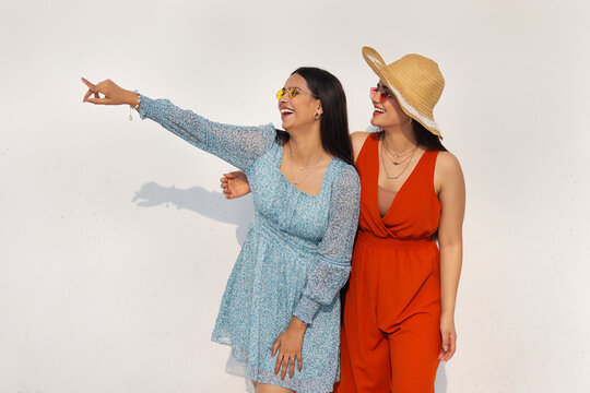 Two smiling young women with sunglasses standing together and pointing against white background