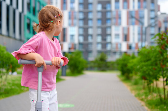 Little girl with a kick scooter looking at modern residential buildings in a city park