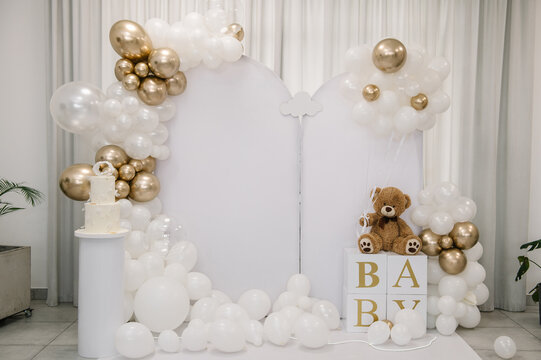Arch. Beautiful photo zone for a birthday, baptism, or baby shower, featuring white arched panels, white and gold balloon garlands, a teddy bear on "BABY" letter blocks, and a cake on a pedestal.