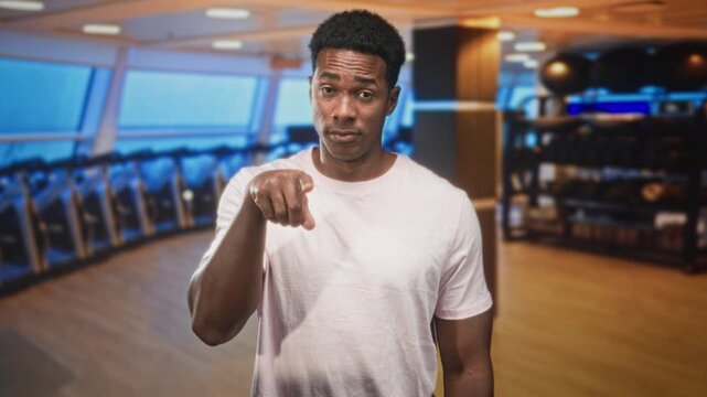 Man points finger at camera in studio gym among treadmills and a weight rack while wearing a white tshirt; challenge accountability.