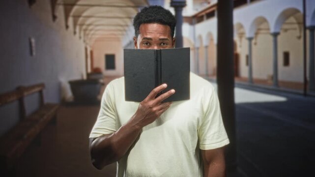 Man peeks over book with hand covering lower face in an arched building corridor; curiosity study focus.