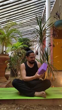 Man wearing black sportswear teaching the use of a purple yoga block on green mat at the garden. instructor blogger filming yoga tips at noon. Vlogger live streaming. User-generated content UGC