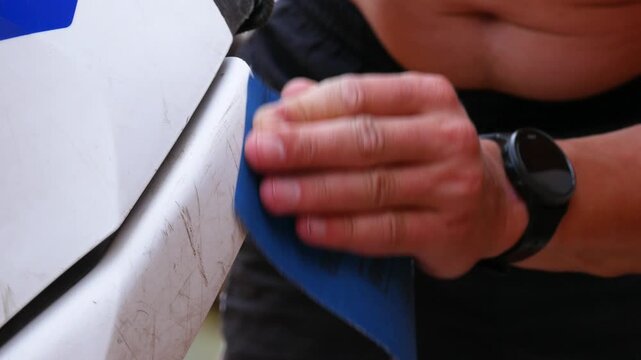 Man sanding car paint surface with abrasive paper. Mechanic repairing scratch on white bumper. Automotive detailing and bodywork maintenance for vehicle restoration.