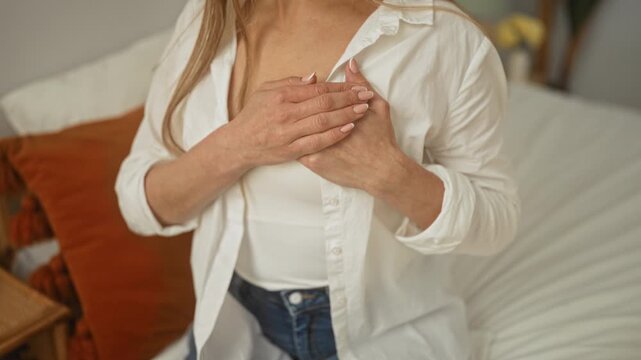 Woman touching hands to chest over white tank top and open shirt while seated on bed in bedroom with pillows and warm light; self care comfort.