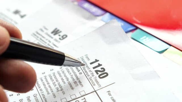 Macro shot of hand holding pen over corporate income tax return form 1120 with colored folders in background