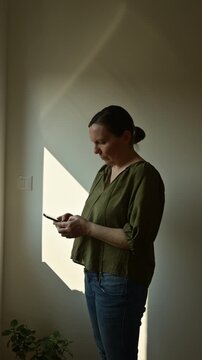 Side profile of middle-aged woman texting on smartphone by window, warm morning sunlight and quiet home lifestyle moment, 4K vertical video