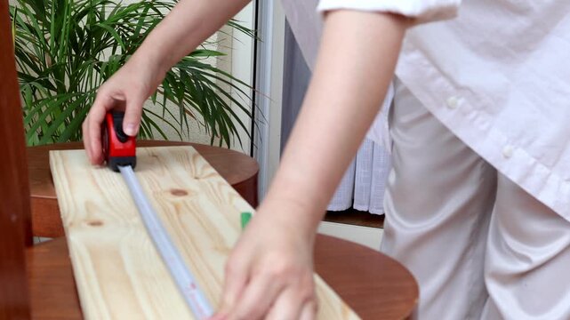 Woman marks cutting lines on a wooden board using a measuring tape and pencil before woodworking at home. Concept of accuracy, DIY furniture making, home renovation and practical craftsmanship.