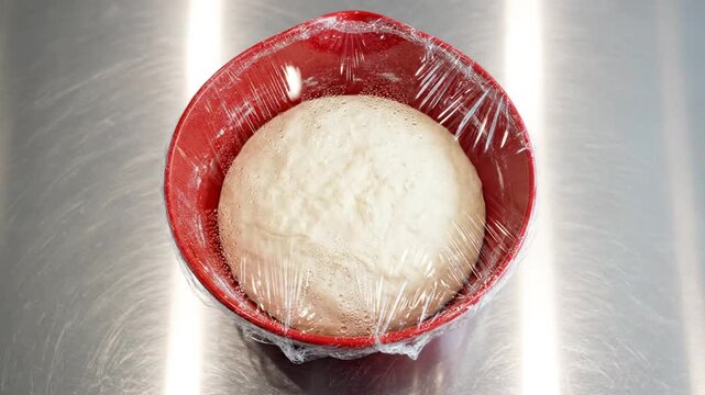 Rising bread dough in a red bowl covered with plastic wrap, showing fermentation and preparation for baking