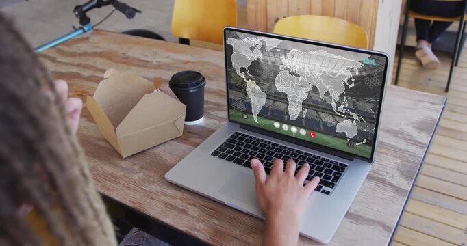 Man working on laptop showing map overlay at cafe while reaching takeout box and eating pastry