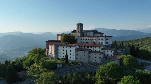Majestic drone shot displays historic sanctuary of Castelmonte on hilltop. Ancient stone fortification walls and religious architecture stand over Friuli region landscape under clear sky.