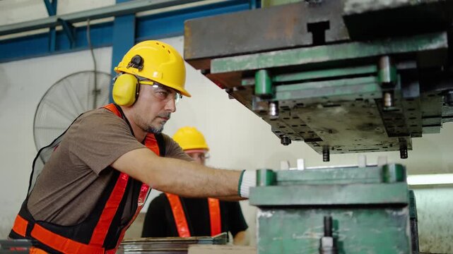 Two male factory workers wearing safety PPE gear operate a heavy industrial hydraulic metal stamping press machine to manufacture and inspect precision sheet metal parts inside a manufacturing plant.