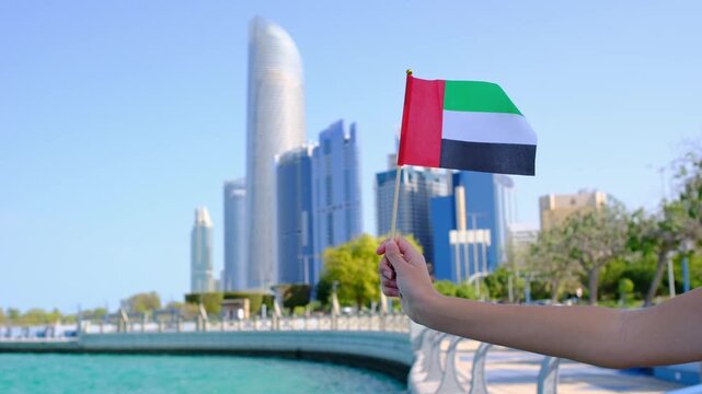 Female hand holding small UAE flag against Abu Dhabi skyline. Symbol of national pride and modernity with iconic skyscrapers in the background showcasing urban elegance and progress