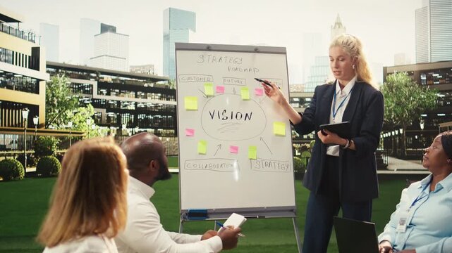 Businesswoman leading whiteboard presentation and explaining ideas to coworkers in outdoor work session, gesturing toward roadmap on board, informal team discussion, for business planning.