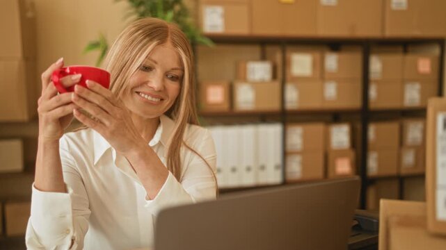 Woman holding red mug with both hands while smiling at laptop amid stacked parcels in small business building; contentment.