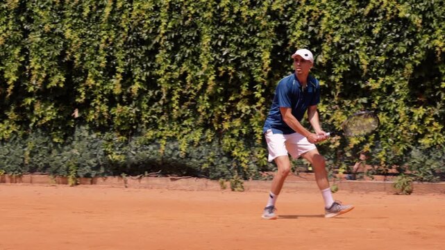Male tennis player performing backhand stroke on outdoor clay tennis court. Concept of competitive tennis, athletic technique, focused training, powerful movement and sports performance.