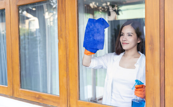 Happy young asian woman wearing gloves cleaning glass windows at home, housekeeper, fresh living environment. Asian girl housewife holding spray sponge cleaning hygiene service window in living room.