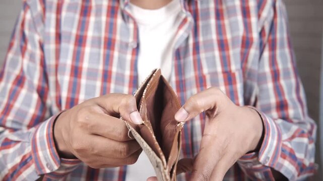A male person in a plaid shirt and white t-shirt, holds an empty wallet with his hands, expressing financial problems, financial crisis, or poverty