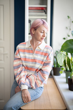 Smiling calm mature woman observing indoor windowsill garden while sitting on table beside window, relaxed carefree female looking at houseplants, tranquil positive lady gazing distracted by thoughts.