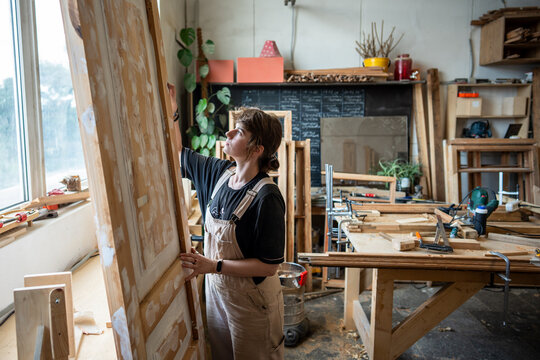 Craftswoman carpenter attentively preparing antique wooden door for final restoration stage, focused female woodworker checking door surface, quality of putty and sanding, handmade craft in studio