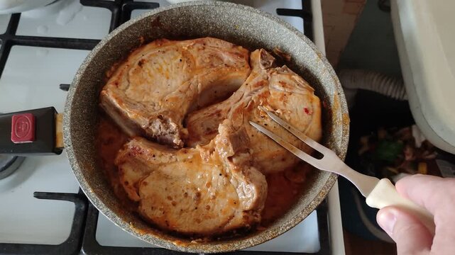 Frying pork chops in a pan with sauce using a fork on a gas stove, top view