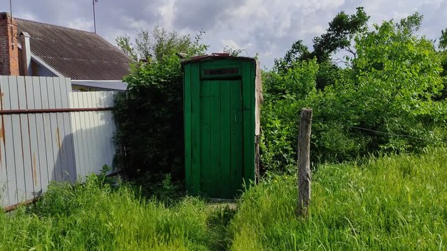 Green wooden outdoor toilet surrounded by tall grass and bushes against a cloudy summer sky
