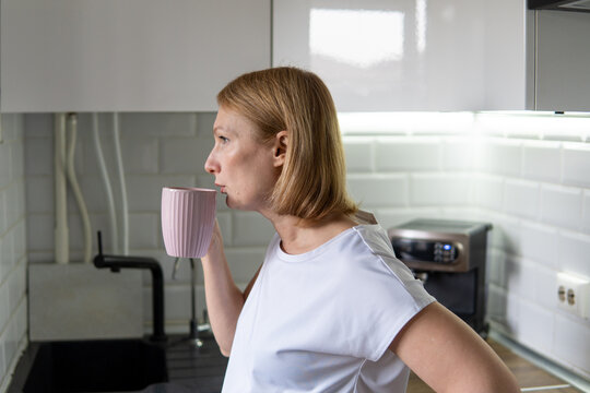 Profile view of woman drinking from pink mug in bright kitchen. Concept of morning coffee.
