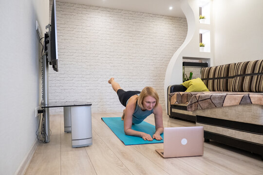 Woman in plank position with leg lift watching online tutorial on laptop. Concept of remote coaching, digital fitness and core strengthening.