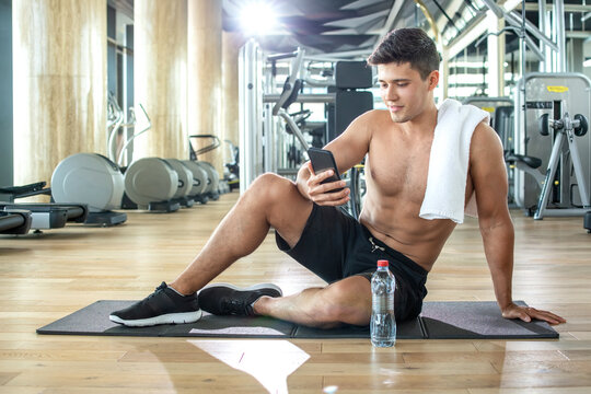 Shirtless fit man resting on a gym mat and checking his smartphone