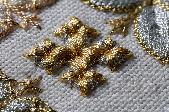 Macro close-up of gold and silver metallic thread embroidery on white linen, photorealistic satin-stitch cross pattern with shallow depth of field