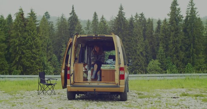 A woman sits comfortably in the back of her camper van, enjoying a peaceful moment surrounded by a lush green forest.