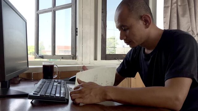A focused adult man wearing a black t-shirt is sitting at his wooden home office desk by the window, writing notes with a pen next to his computer and a glass of black coffee.