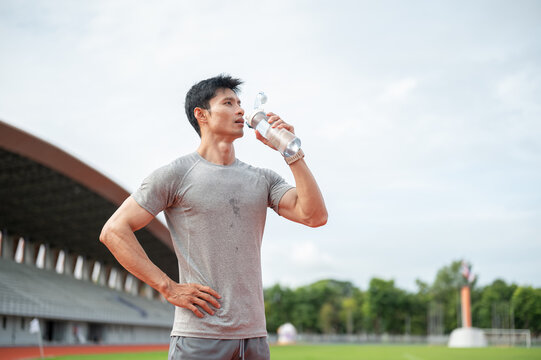 Thirsty Asian man drinking water from bottle while standing on track in sports stadium.