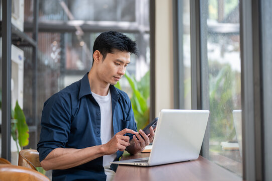 Young Asian entrepreneur using smartphone while working on laptop at table in cafe