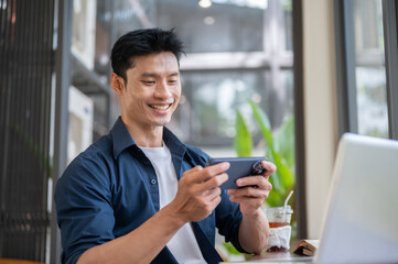 Fototapeta premium Asian man smiling while using or watching media on smartphone at table in modern cafe workspace.